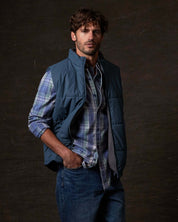 Man wearing a Matte Coated Shell Puffer Vest in Slate Blue over a plaid shirt, showcasing its stylish utility.