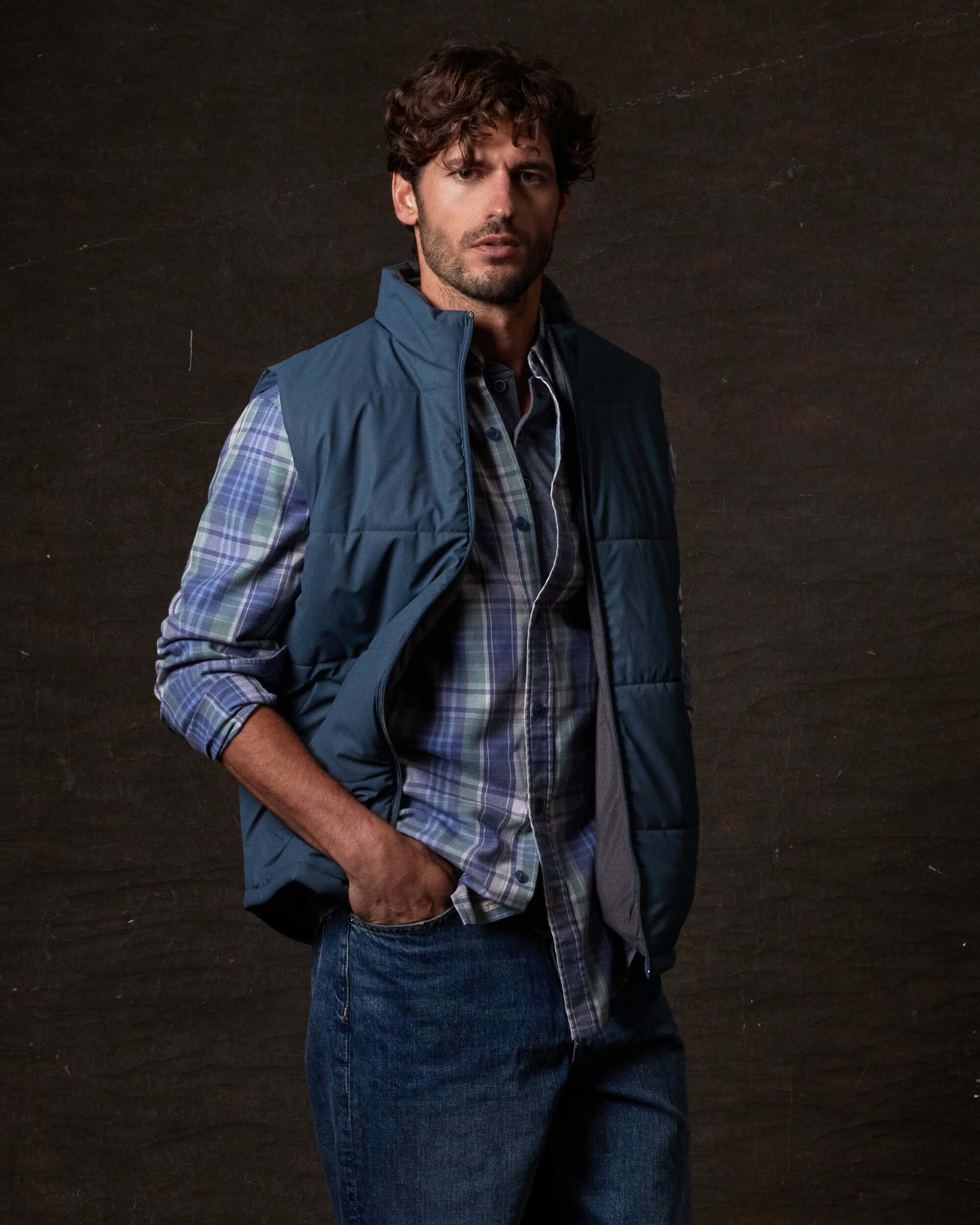 Man wearing a Matte Coated Shell Puffer Vest in Slate Blue over a plaid shirt, showcasing its stylish utility.