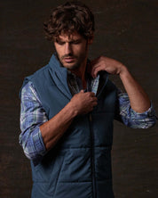 Model wearing Matte Coated Shell Puffer Vest in Slate Blue, adjusting the zip closure, set against a dark background.
