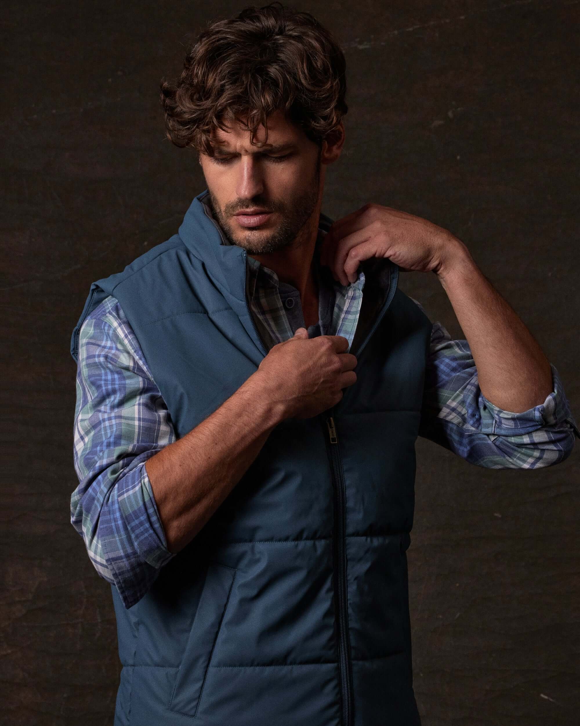 Model wearing Matte Coated Shell Puffer Vest in Slate Blue, adjusting the zip closure, set against a dark background.