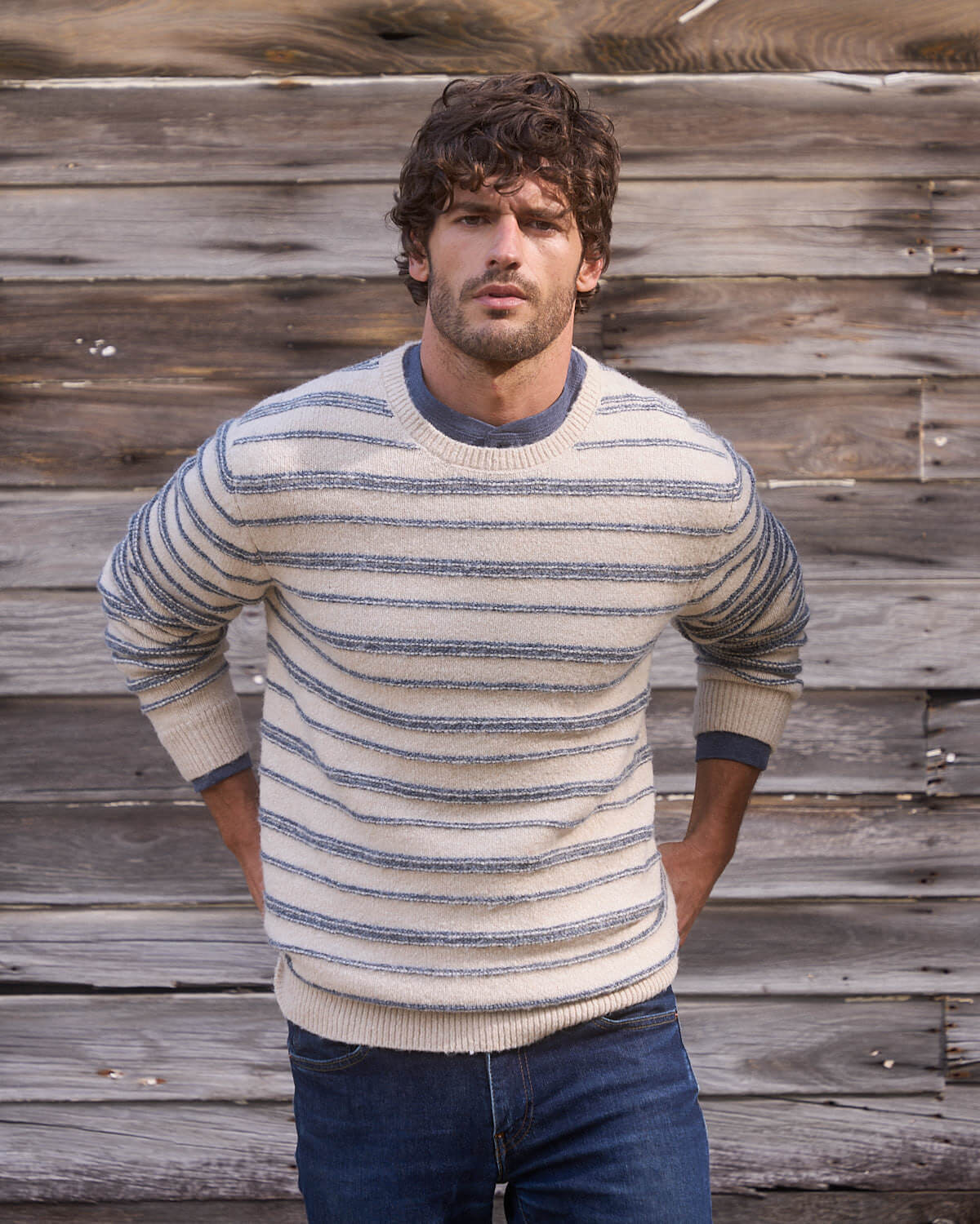 Model wearing a cream and blue fuzzy striped crew sweater, showcasing classic style against a rustic wooden backdrop.