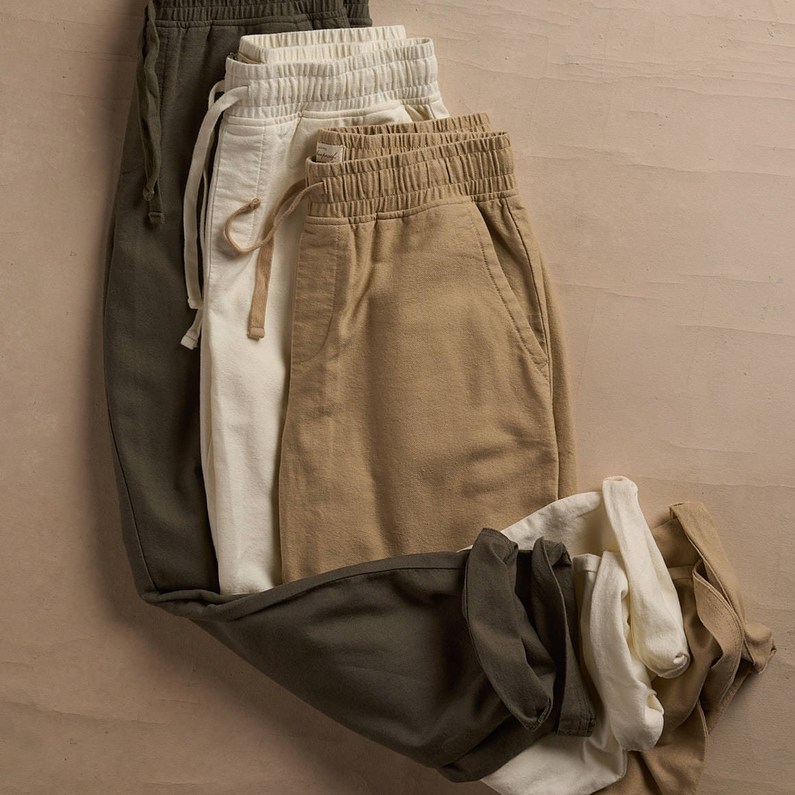 Casual sweatpants in various earth tones: olive, beige, and cream laid out on a textured surface.