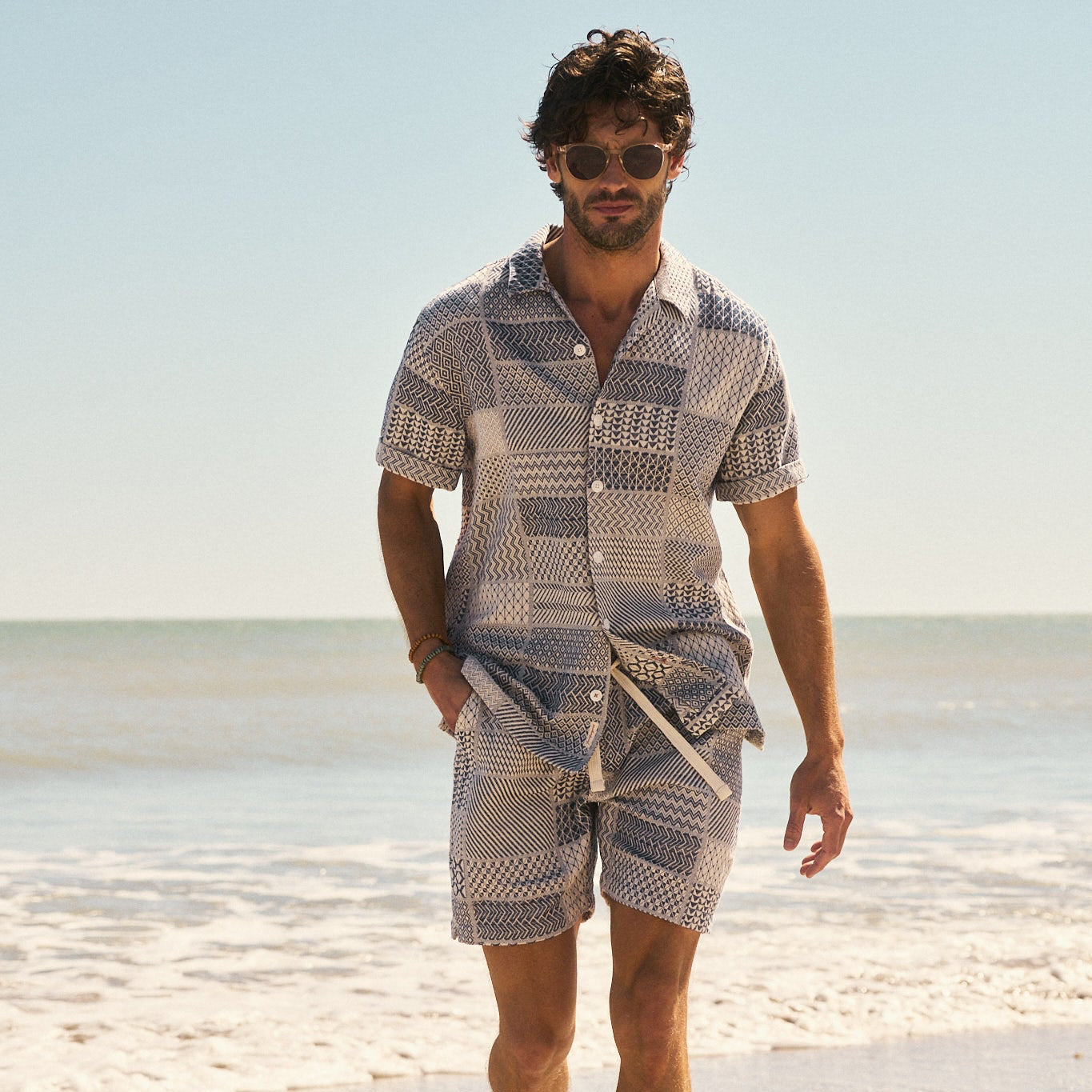 Man in patterned shirt and shorts walking along the beach with sunny skies and ocean waves in the background.