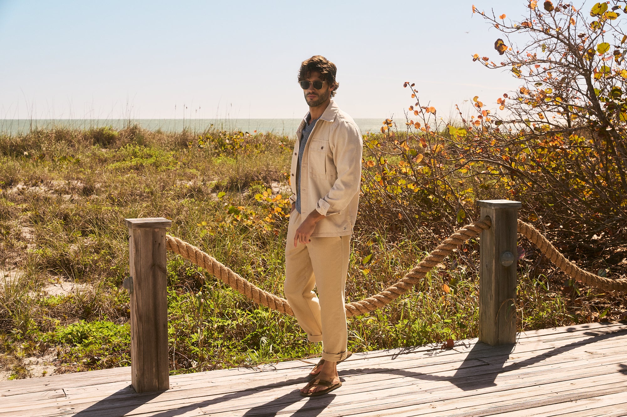 A stylish man walks along a wooden path by the beach, wearing a light jacket and casual pants under clear skies.