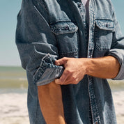 Denim Shirt In Acid Blue styled with rolled-up sleeves at the beach, showcasing its soft fabric and rugged look.