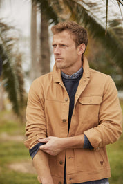 Man in a brown jacket standing outdoors among palm trees, looking thoughtfully into the distance.