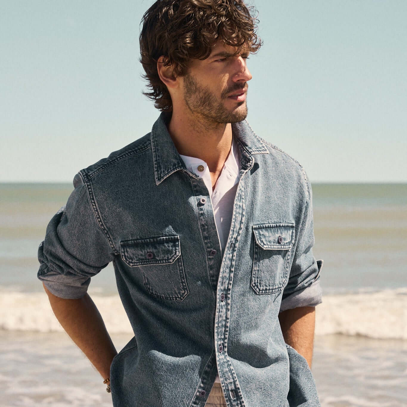 Man wearing Denim Shirt In Acid Blue, standing on the beach with ocean in the background.