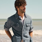 Man wearing Denim Shirt In Acid Blue, standing on the beach with ocean in the background.