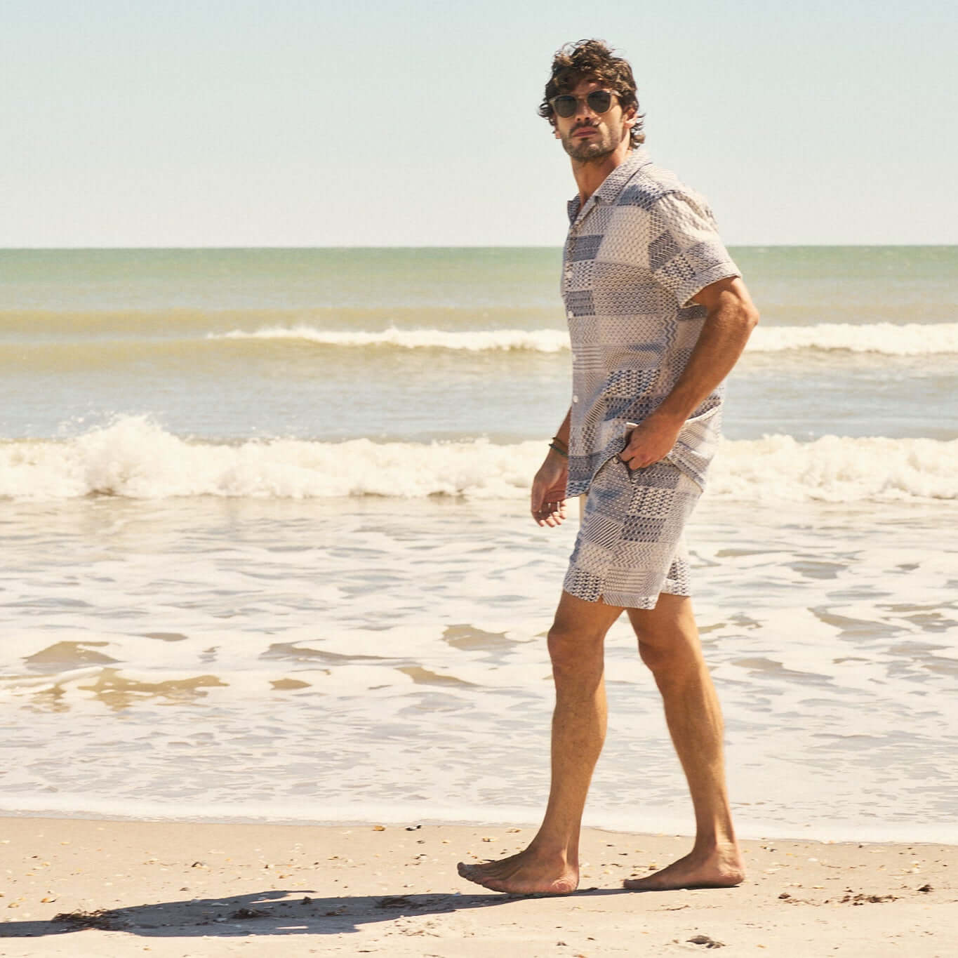 Model wearing a cream and navy jacquard resort shirt and matching shorts by the beach, embodying retro vacation style.