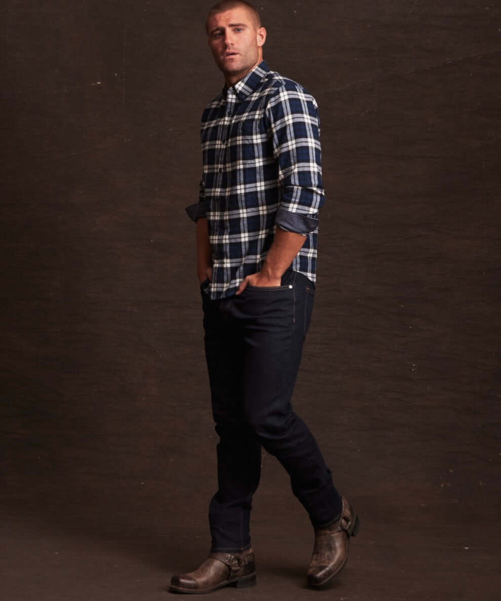 Man wearing a long sleeve brushed flannel shirt in blue plaid, paired with dark jeans and boots, set against a dark background.