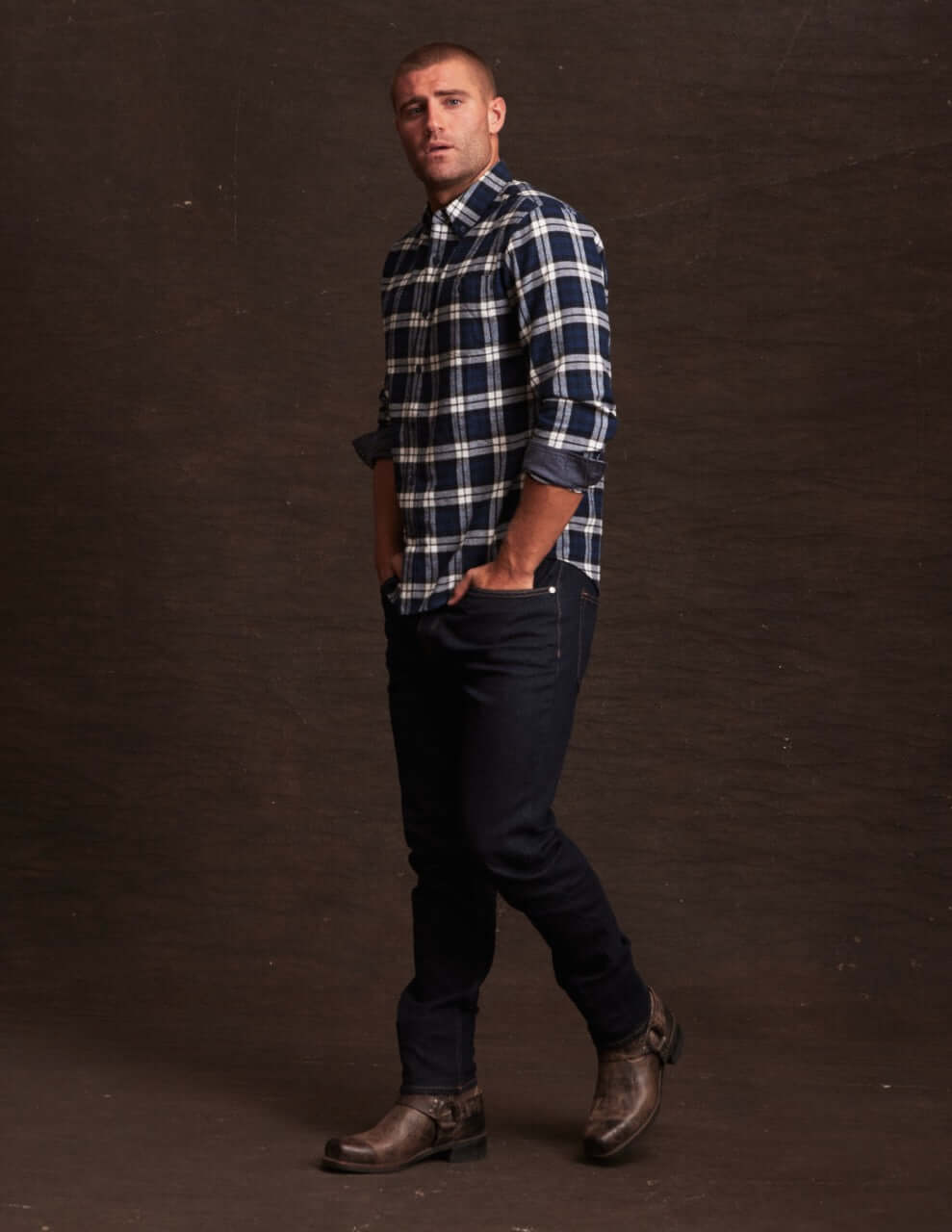 Man wearing a long sleeve brushed flannel shirt in blue plaid, paired with dark jeans and boots, set against a dark background.