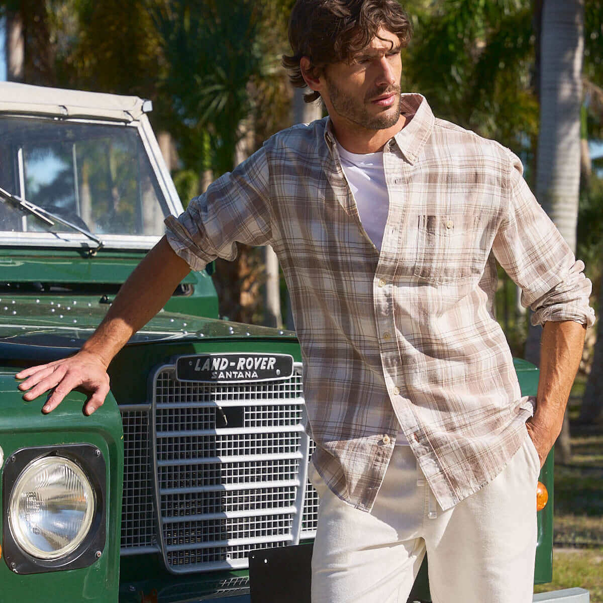 Model wearing Long Sleeve Faux Linen Plaid Shirt in Natural, standing next to classic Land Rover in sunny outdoor setting.