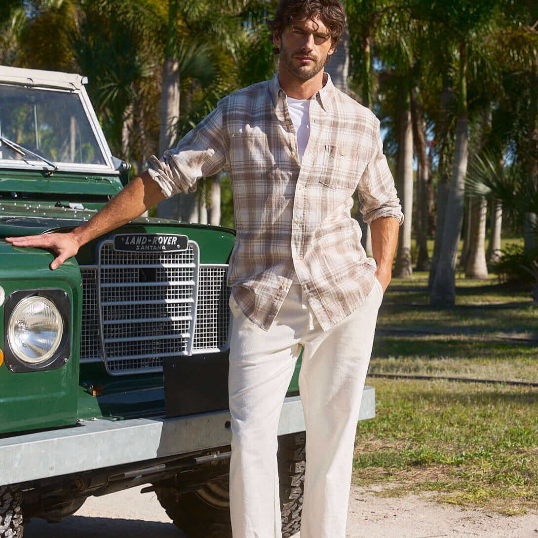 Man wearing Long Sleeve Faux Linen Plaid Button Down shirt in Natural beside a classic green Land Rover in a sunny outdoor setting.