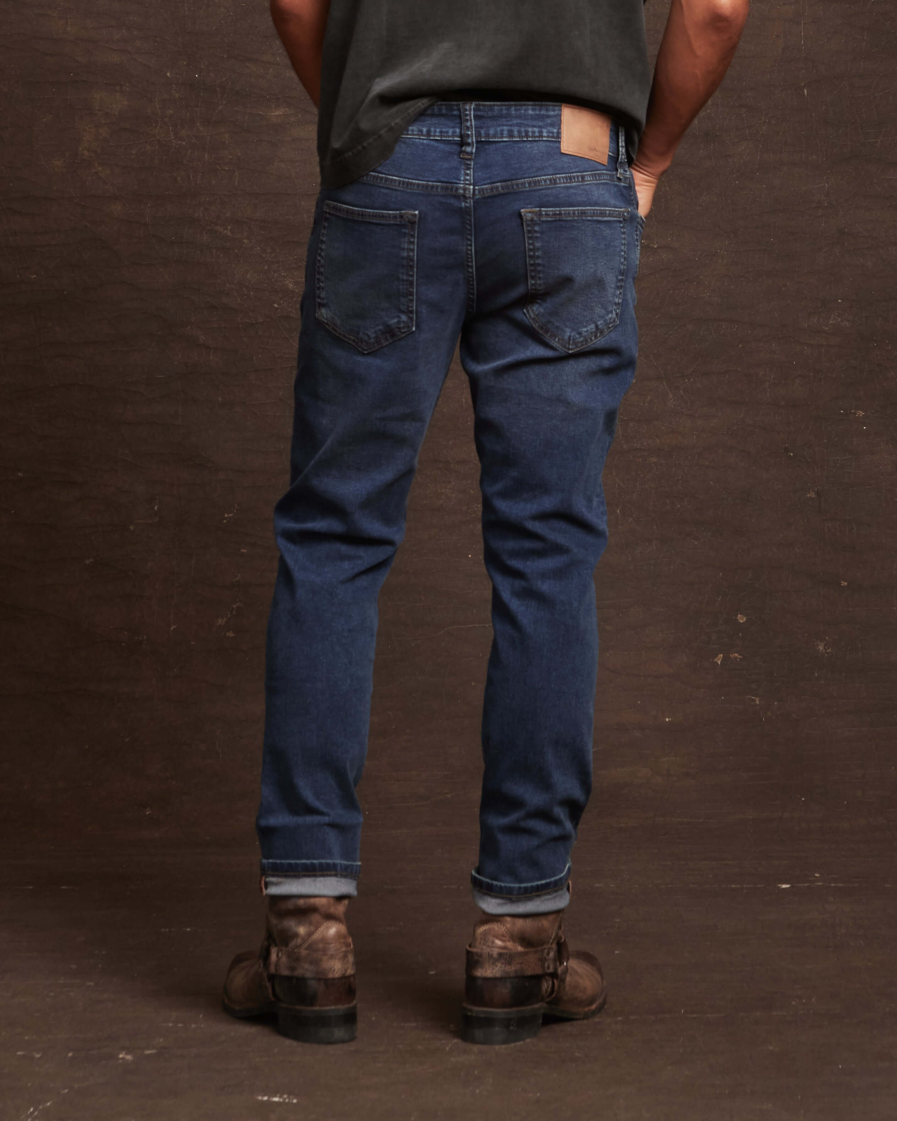 Back view of men's slim fit denim jeans in soft blue with rolled cuffs and brown boots against a textured background.