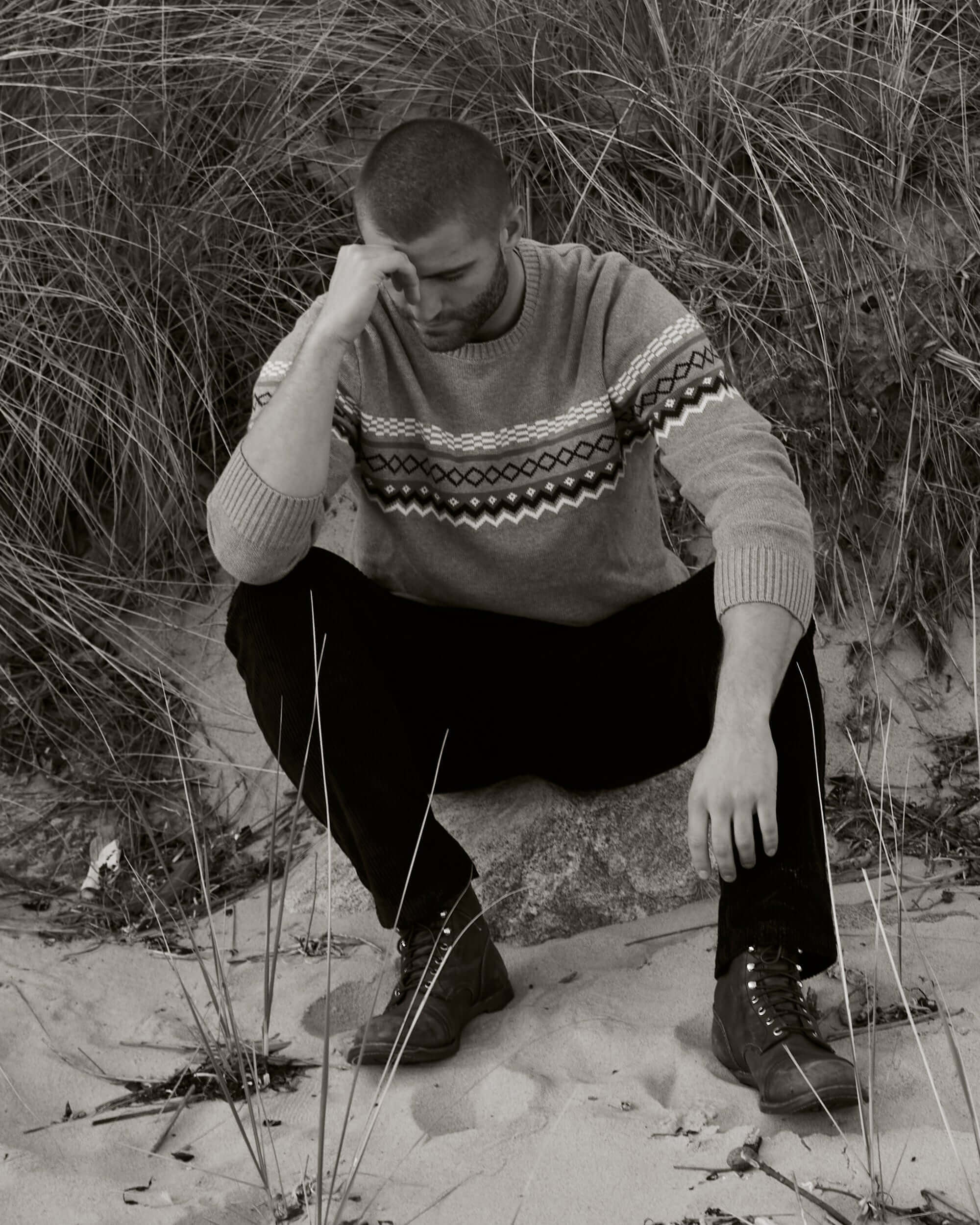 Man in pale grey Nordic crewneck sweater sitting on sand, surrounded by grass. Demonstrates stylish comfort in outdoor setting.