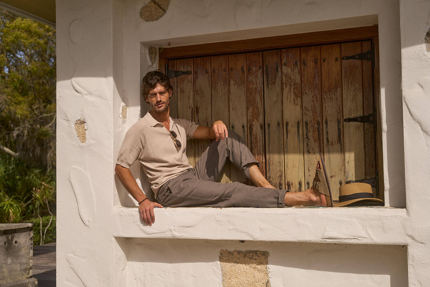 Man lounging in an open collar polo sweater, showcasing a relaxed style against a rustic window backdrop.