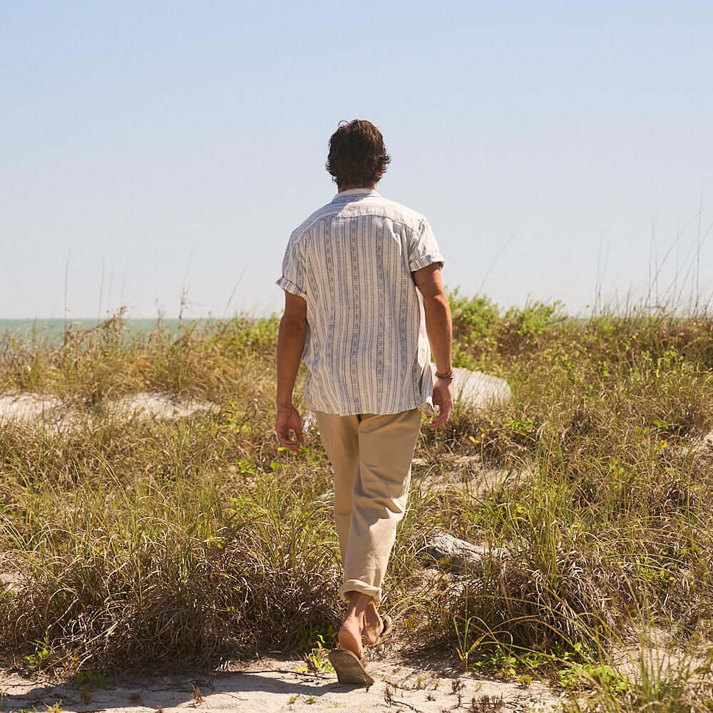 Man in Short Sleeve Aztec Dobby Shirt walking along a sandy path by the beach, showcasing a classic and comfortable style.