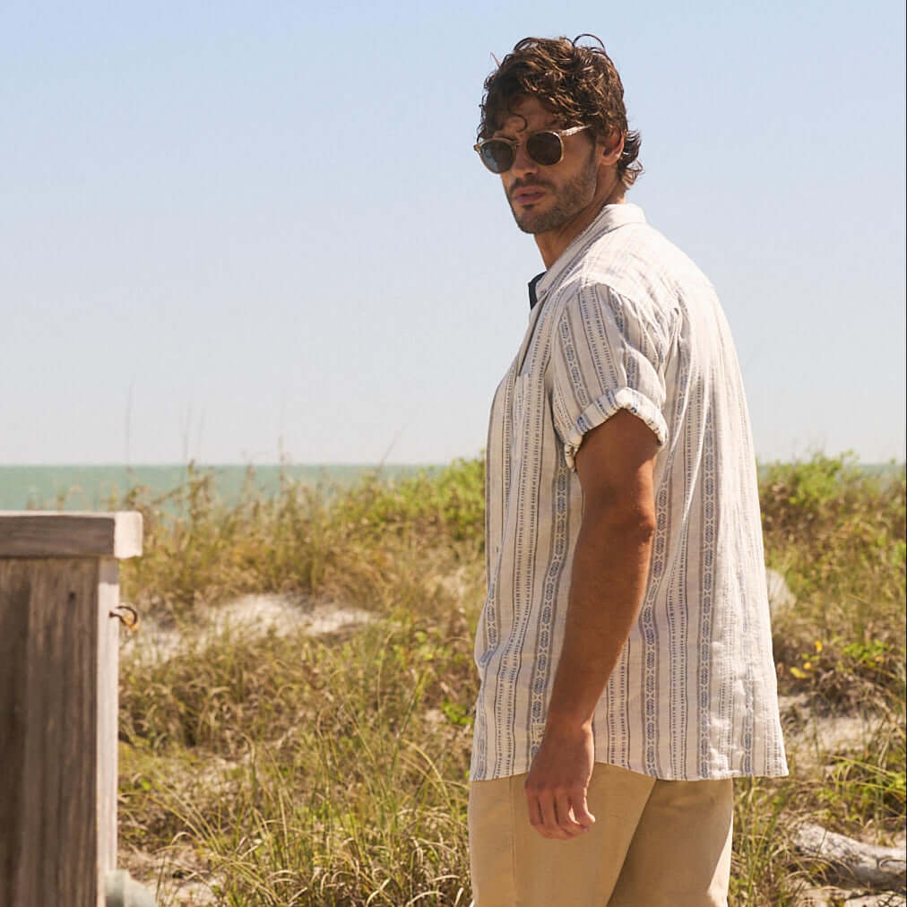 Casually styled man in sunglasses and patterned shirt standing on a beach with grass and ocean backdrop.
