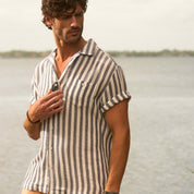 Model wearing a Short Sleeve Cotton Linen Airtex Stripe Shirt in Insignia Blue by the water, showcasing vacation style.