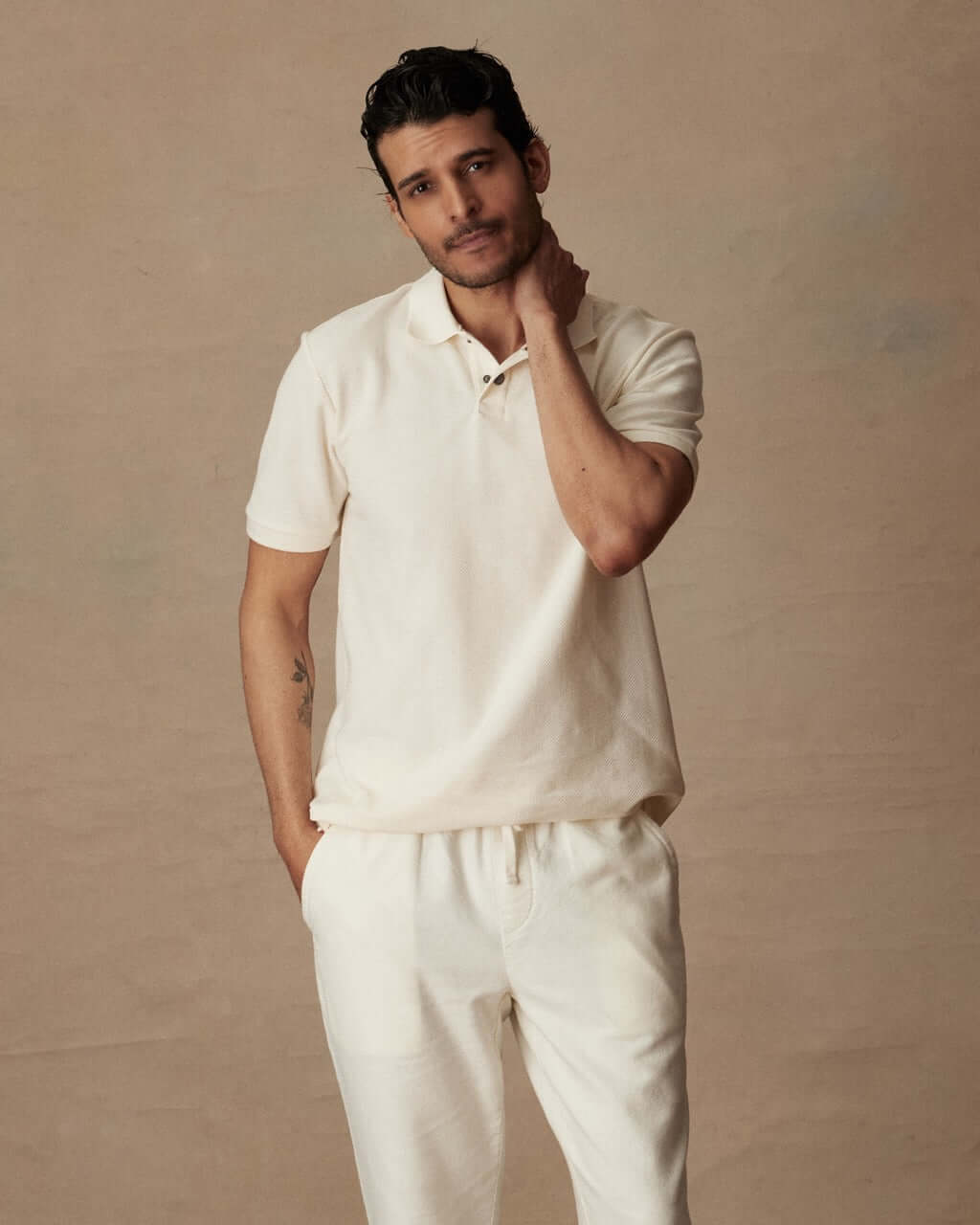 Man in short sleeve ivory honeycomb pique polo shirt and cream pants on neutral backdrop.