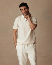 Man in short sleeve ivory honeycomb pique polo shirt and cream pants on neutral backdrop.