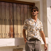 Man wearing a Short Sleeve Linen Cotton Camp Shirt in Sugar Almond by a rustic wooden door, showcasing its bold print and breezy style.