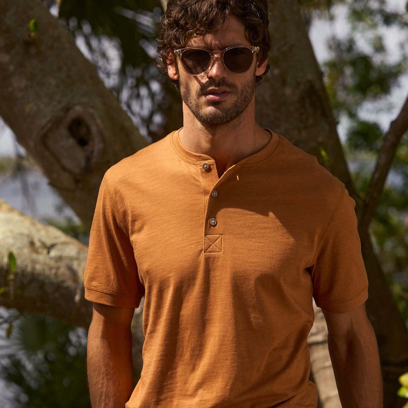 Man in sunglasses wearing a short sleeve melange henley shirt in adobe color, set against a natural background.