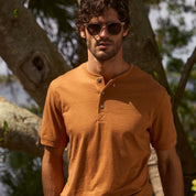 Man in sunglasses wearing a short sleeve melange henley shirt in adobe color, set against a natural background.