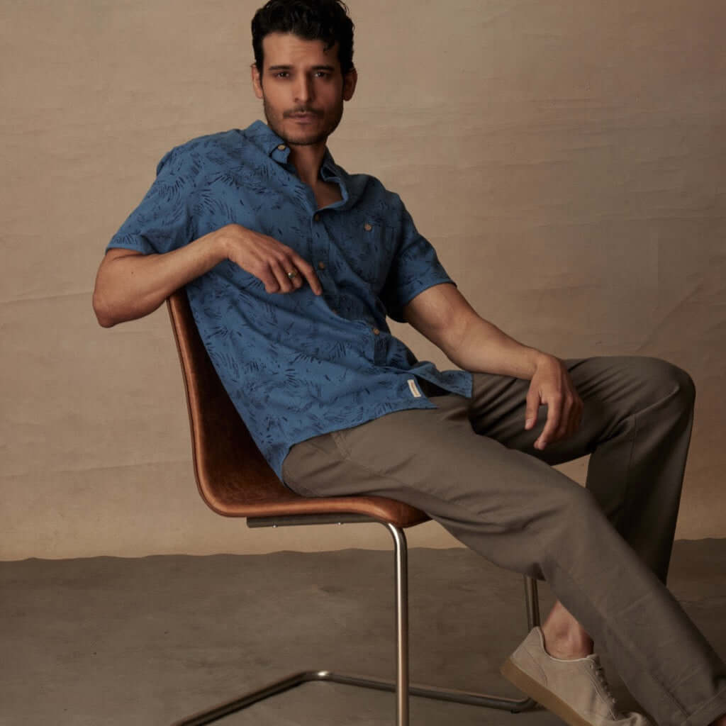Model in a blue ashes short sleeve print linen cotton shirt, seated in a modern chair, showcasing a relaxed fit and abstract patterns.