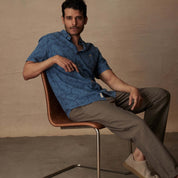 Model in a blue ashes short sleeve print linen cotton shirt, seated in a modern chair, showcasing a relaxed fit and abstract patterns.