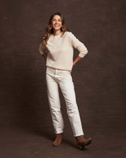 Smiling woman wearing a women's cashmere long sleeve sweater in ecru with white jeans and brown ankle boots.