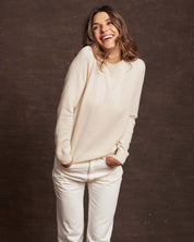 Smiling woman wearing a women's ecru cashmere long sleeve sweater paired with white pants.