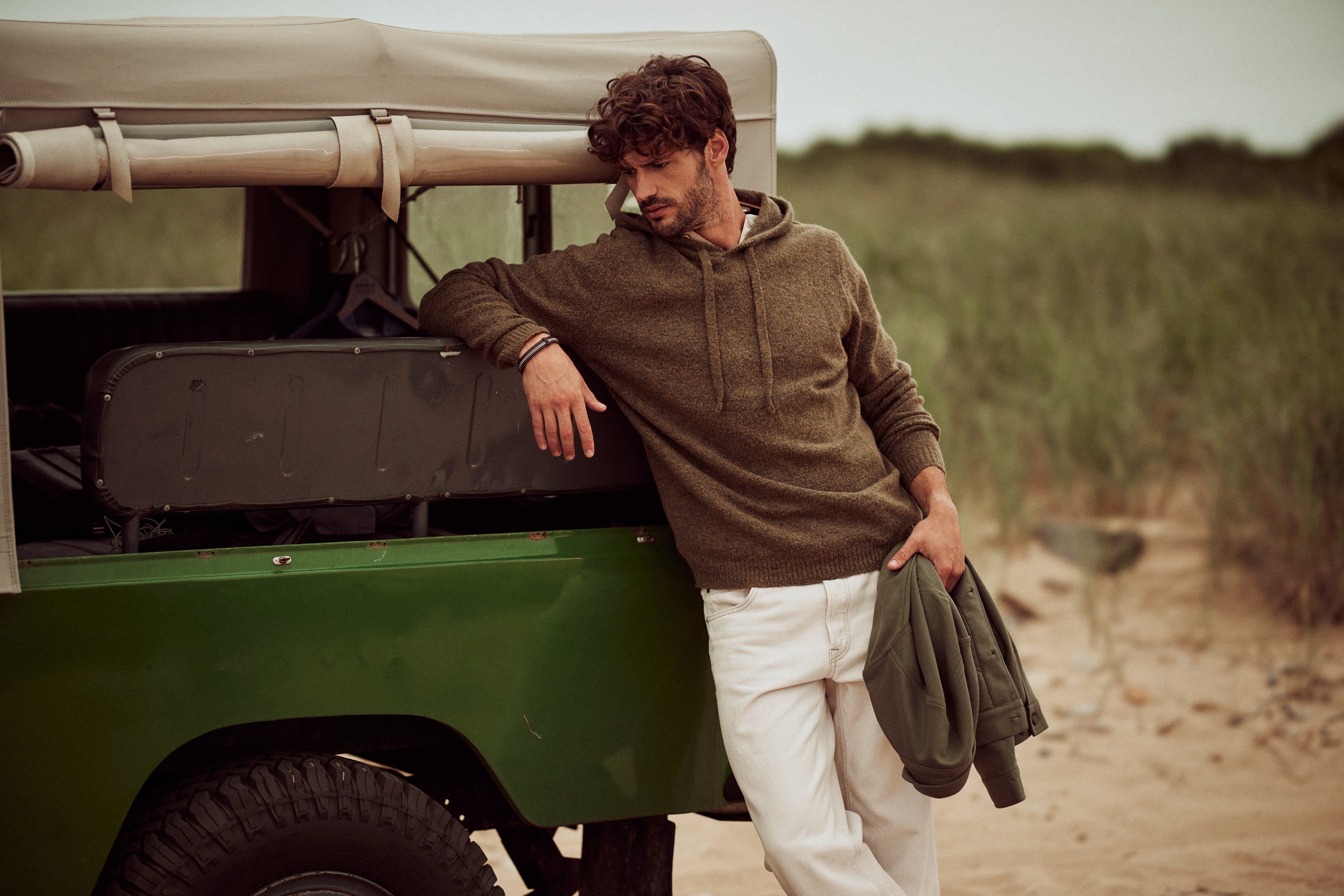 Boucle Hoodie Sweater in Moss worn by a man leaning against a vintage green jeep in a sandy outdoor setting.