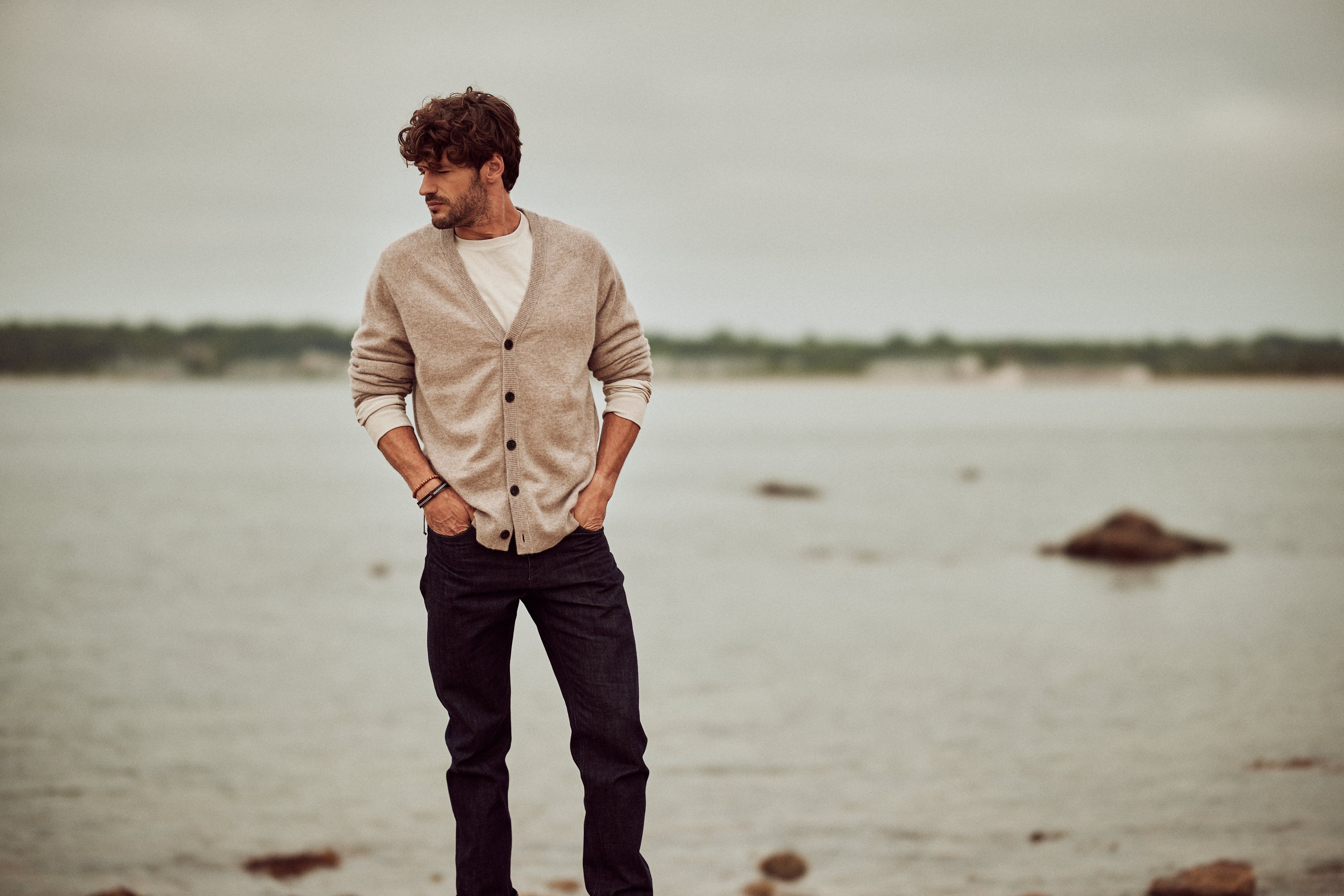 CASHMERE BUTTON CARDIGAN in TAUPE worn by a man standing by the water, showcasing a stylish and comfortable look.