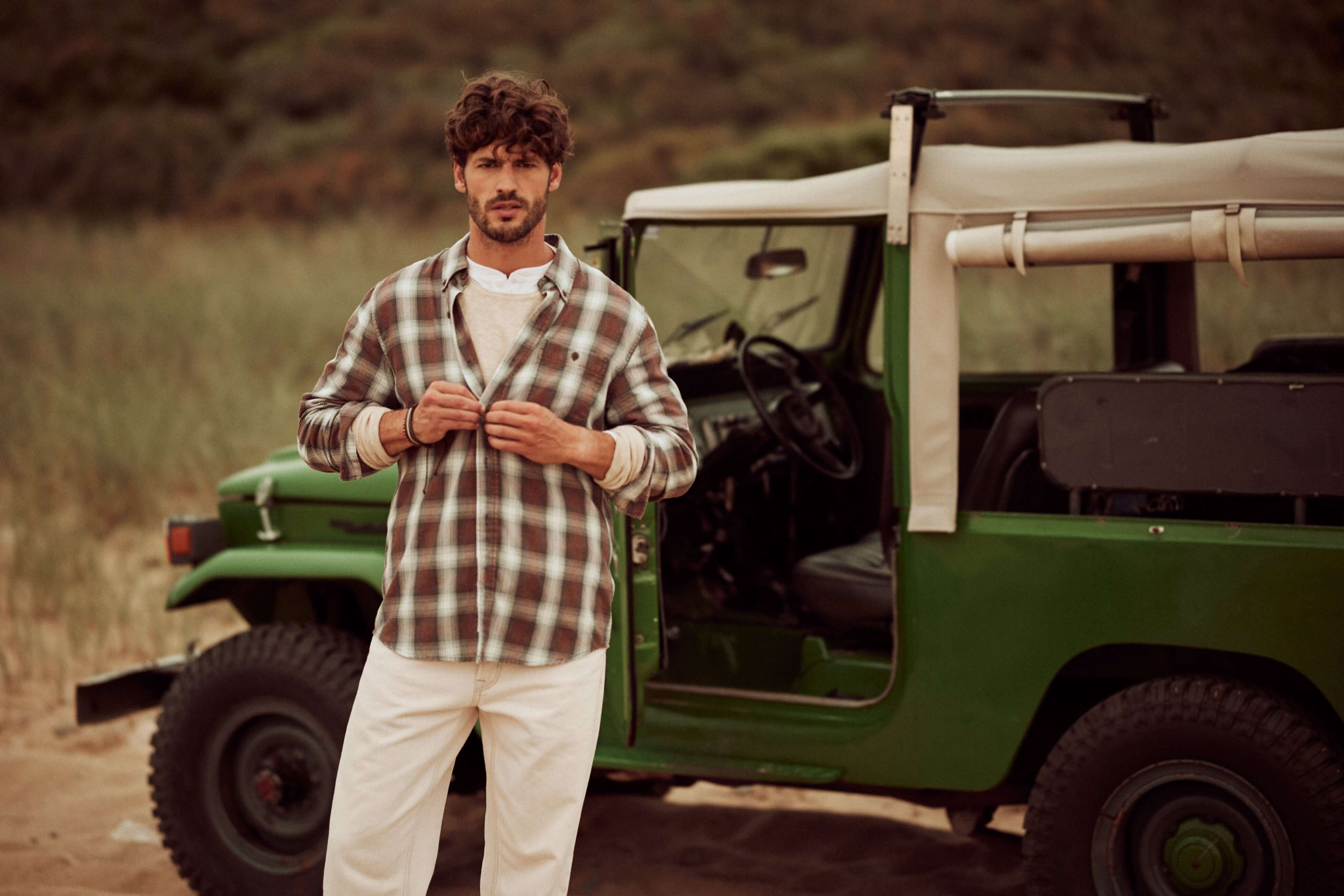 Man wearing a long sleeve burnout flannel shirt with a weathered look, standing near a vintage green jeep on the beach.