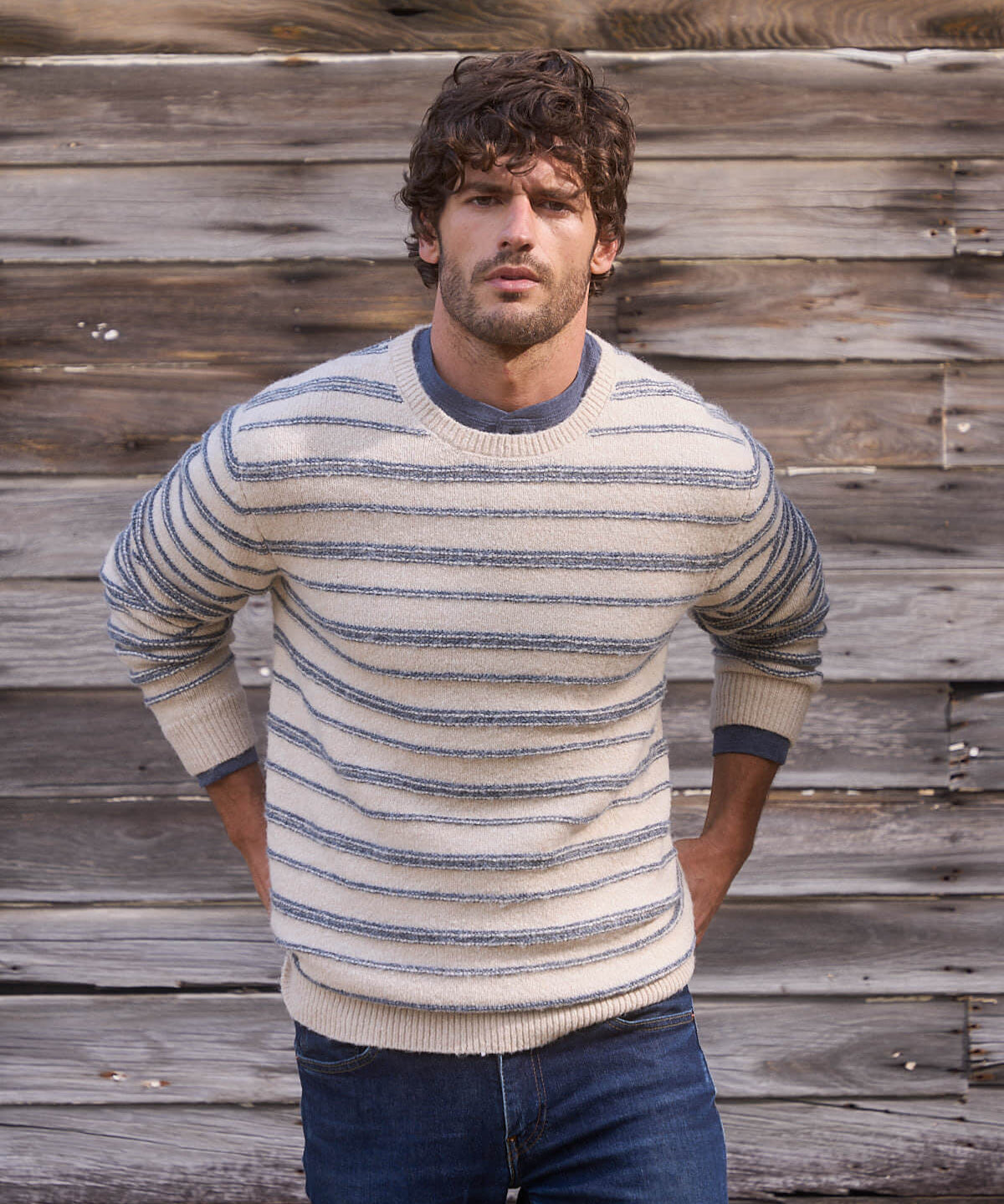 Model wearing a cream and blue fuzzy striped crew sweater, showcasing classic style against a rustic wooden backdrop.