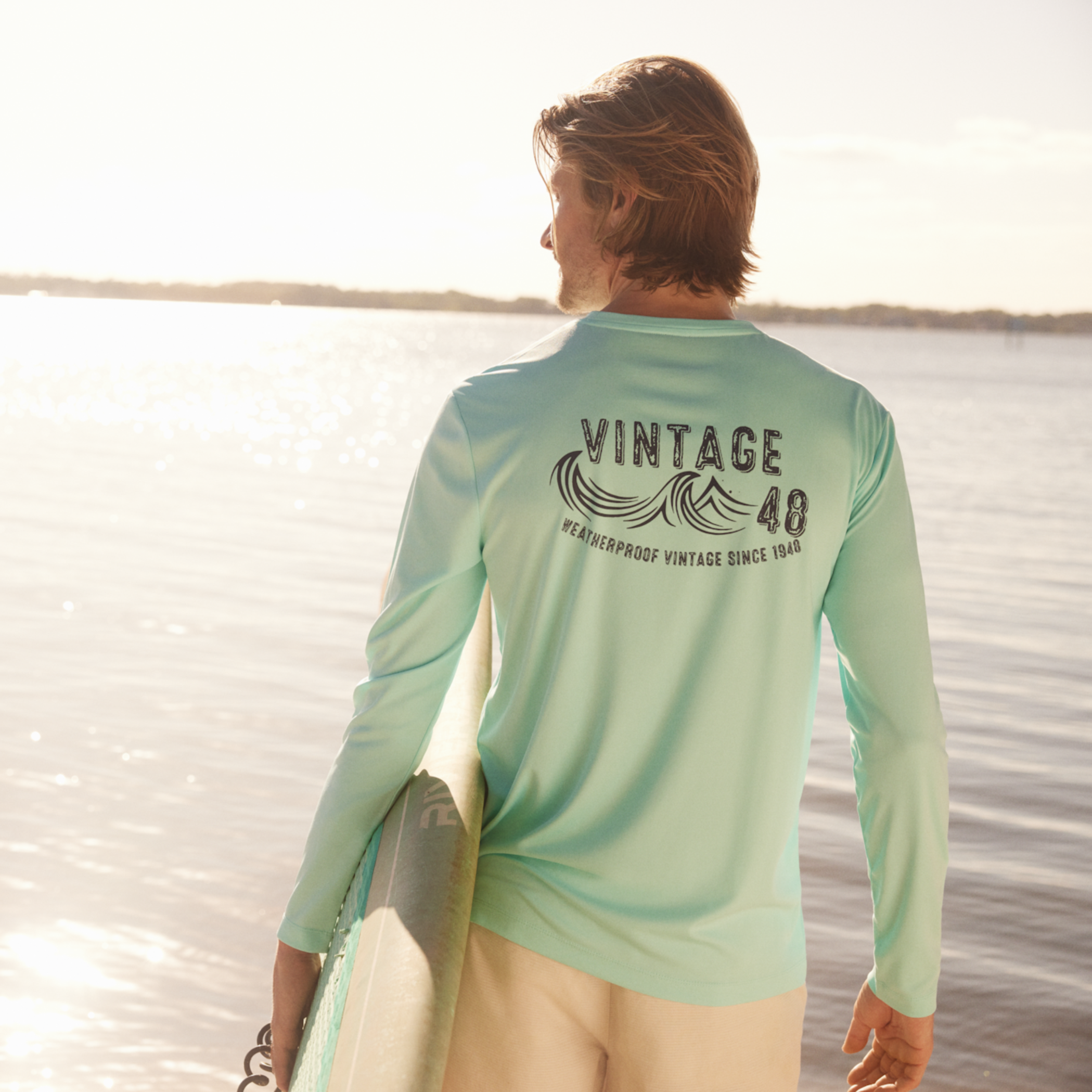 Long Sleeve Sun Shirt Spf In Aruba Blue worn by a man by the water, displaying vintage graphics and sun protection.