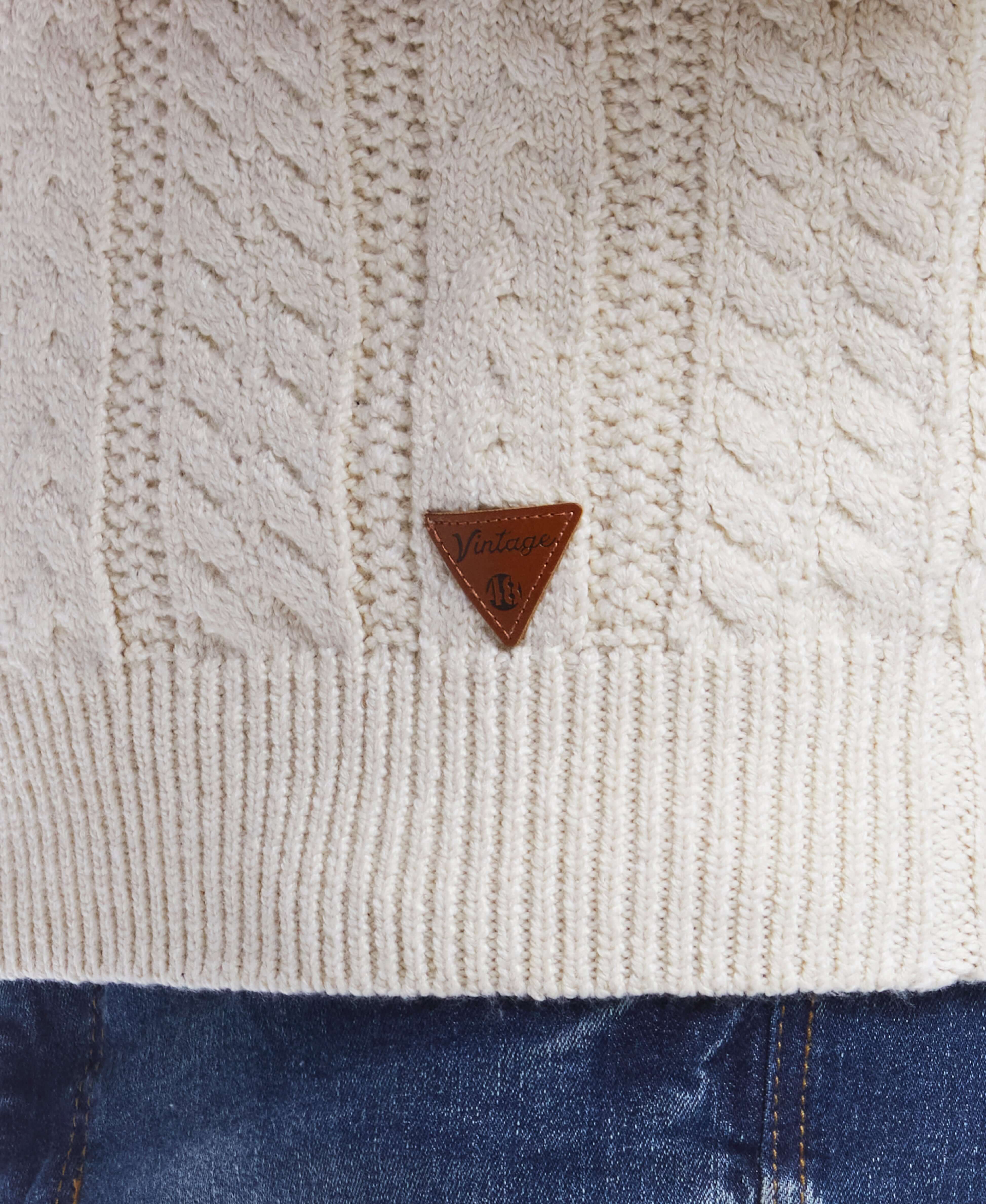 Close-up of white allover cable knit crew sweater with leather logo patch, perfect mid-layer for cooler weather.