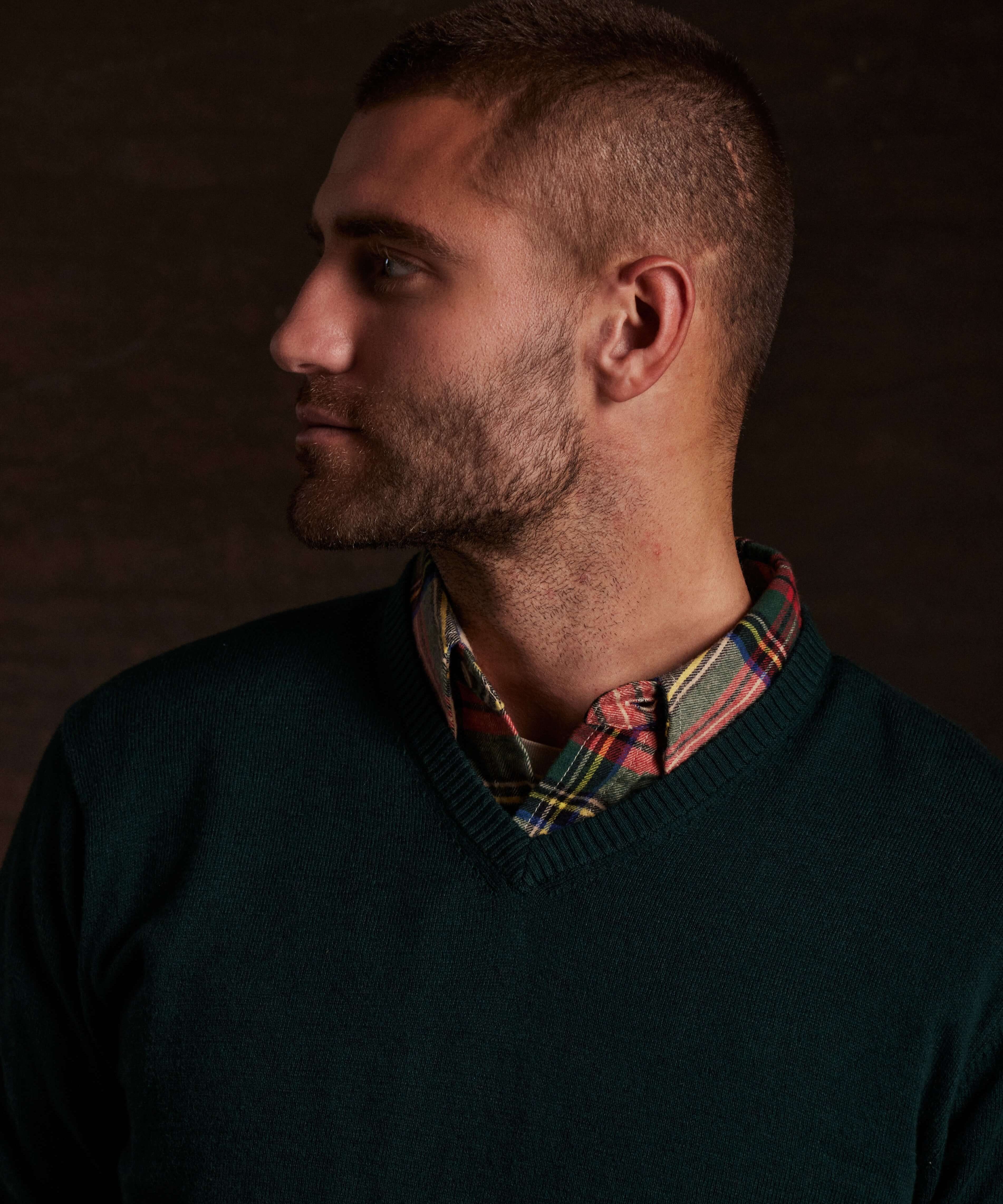 Model showcasing a cotton cashmere v-neck sweater in pine, layered over a plaid shirt.