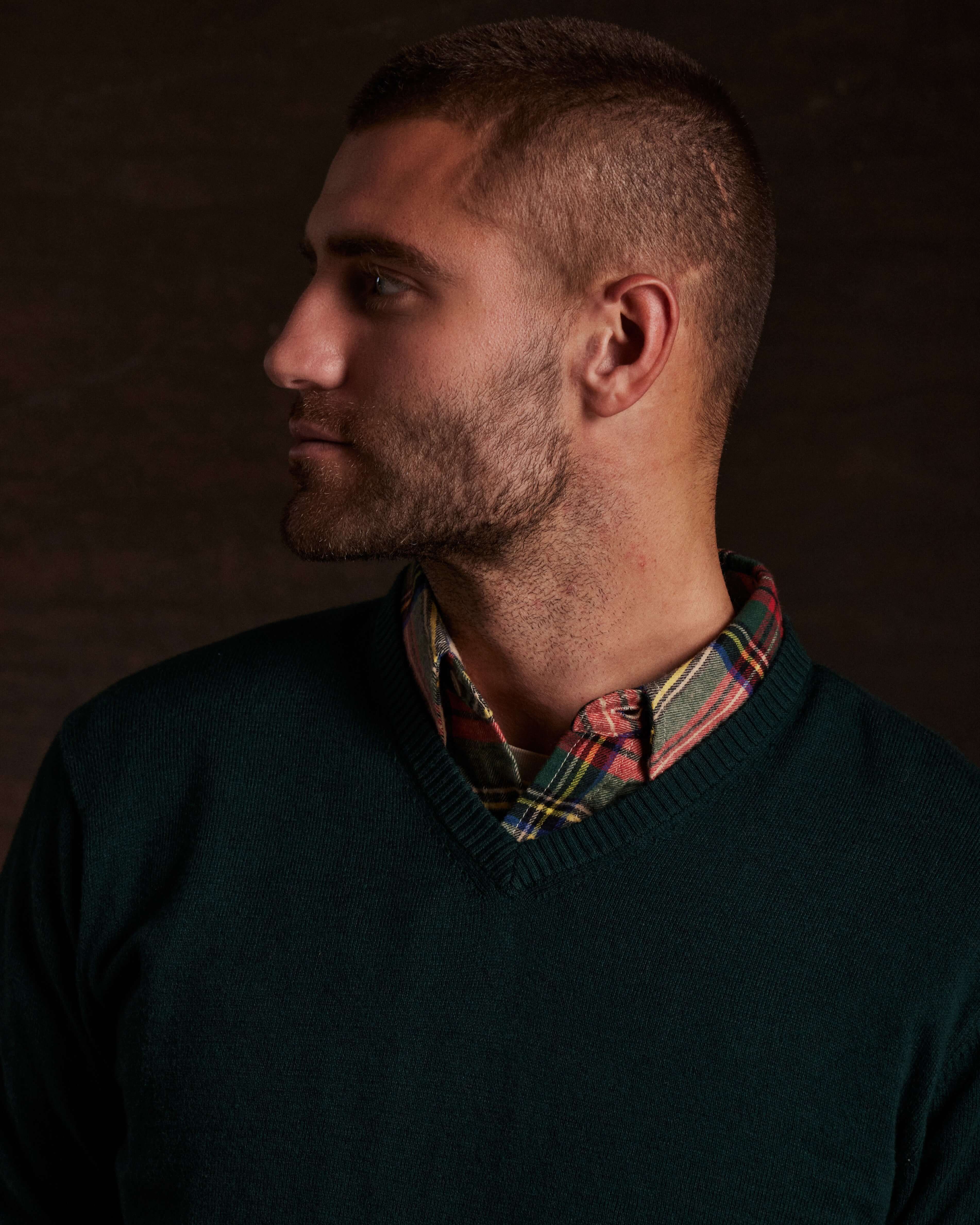 Model showcasing a cotton cashmere v-neck sweater in pine, layered over a plaid shirt.