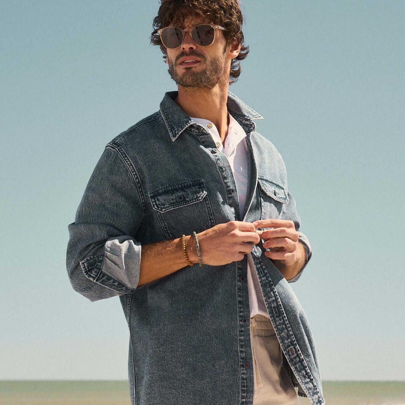 Stylish man in sunglasses wearing a denim jacket by the beach, embracing a relaxed summer vibe.