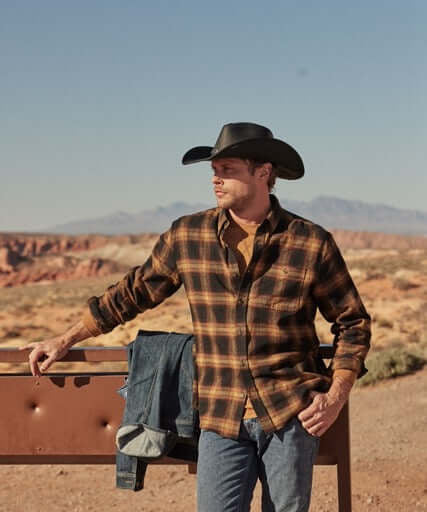 Man wearing Long Sleeve Brushed Flannel in Inca Gold, styled with a cowboy hat, against a rugged outdoor backdrop.