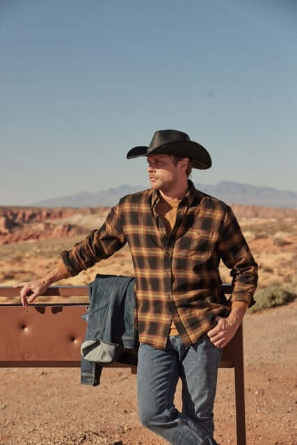 Man wearing Long Sleeve Brushed Flannel in Inca Gold, styled with a cowboy hat, against a rugged outdoor backdrop.