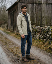 Poly Memory Safari Jacket with Hood in Sea Grass worn by a man standing on a gravel path, surrounded by rustic scenery.