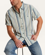 Short Sleeve Seersucker Stripe in Green Slate worn by a model sitting on a chair, showcasing its vertical stripe pattern.