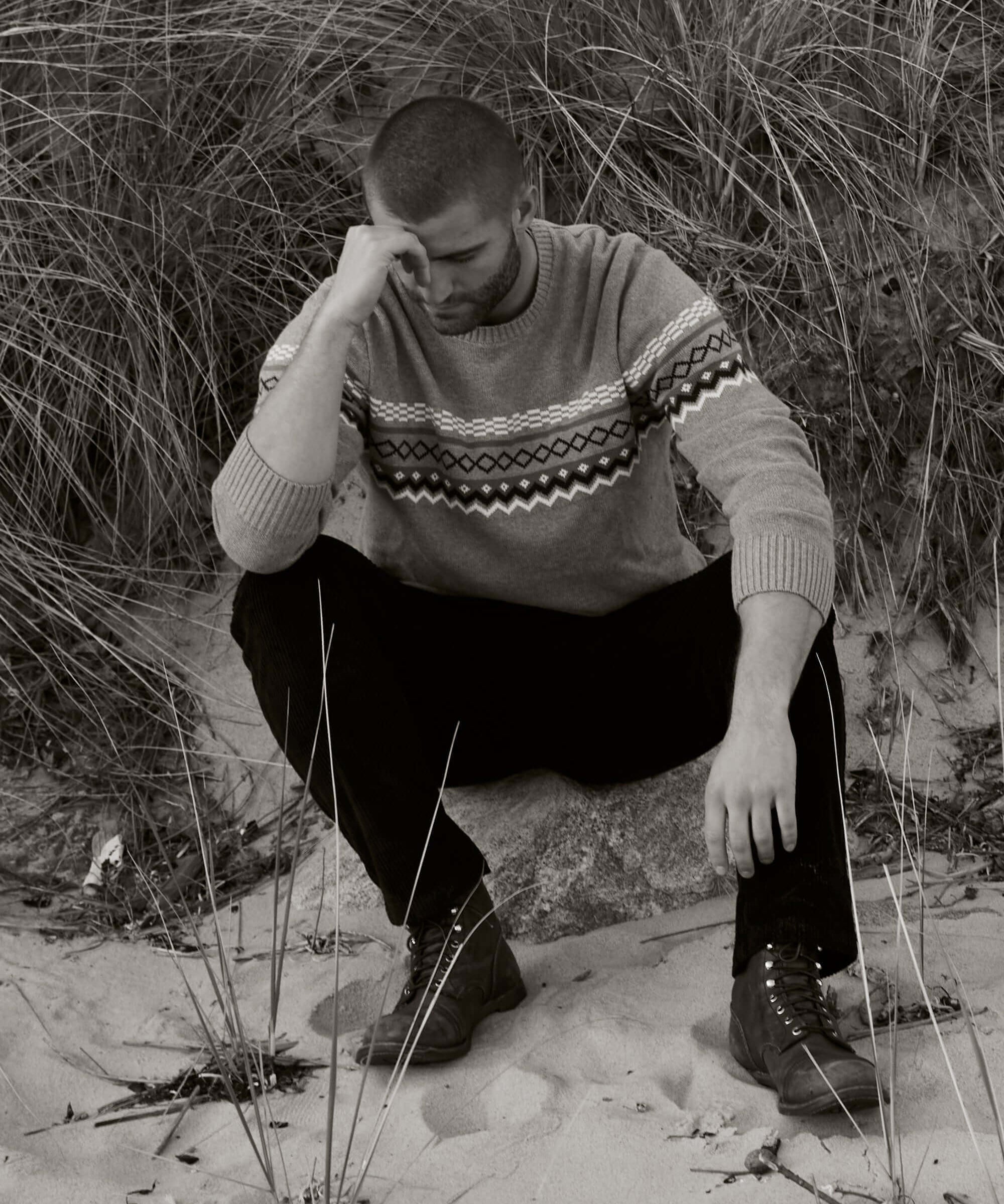 Man in pale grey Nordic crewneck sweater sitting on sand, surrounded by grass. Demonstrates stylish comfort in outdoor setting.