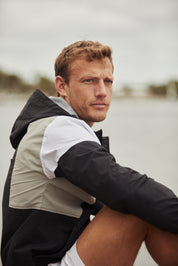 Contemplative man in sporty jacket sitting by water, reflecting on a cloudy day.