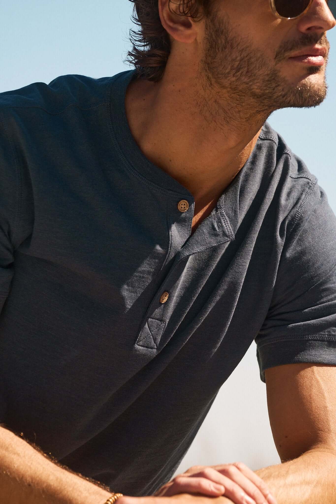 Man wearing a stylish blue henley shirt, exuding a casual yet sophisticated vibe in a sunlit outdoor setting.