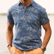 Short sleeve printed polo shirt in dark indigo with tropical palm design on a model by the water.