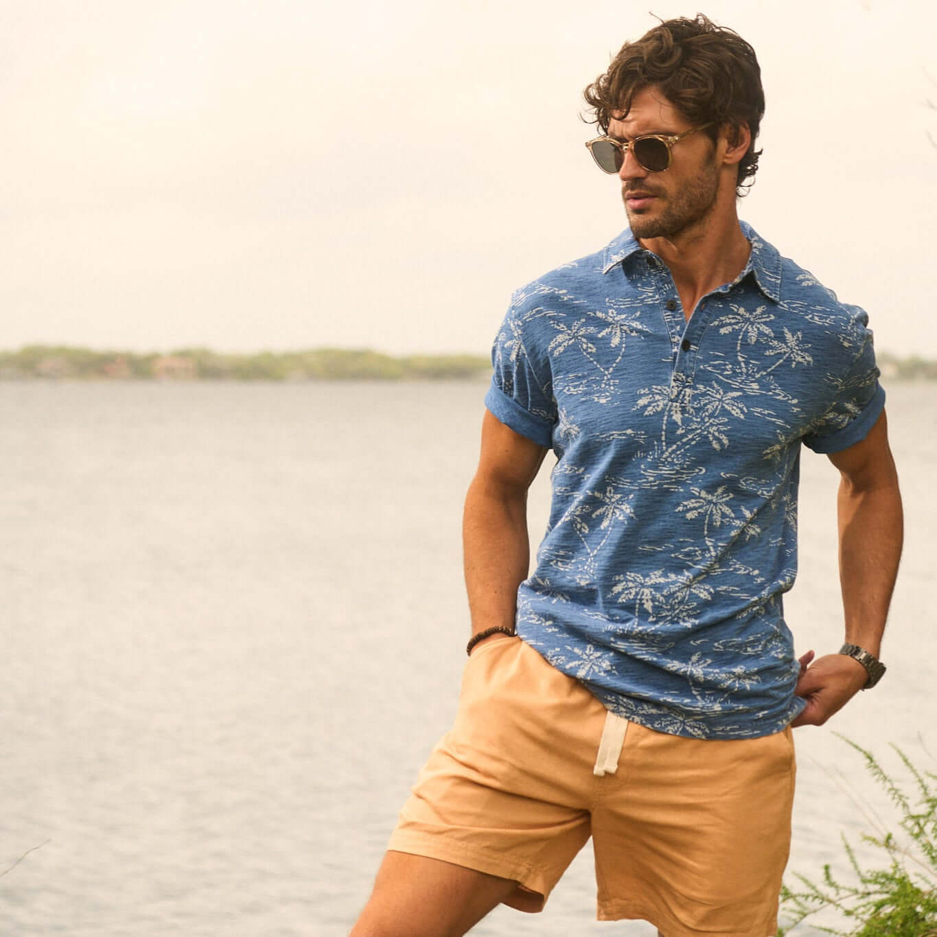 Man in dark indigo short sleeve printed polo shirt by water, showcasing vintage tropical print and casual summer style.